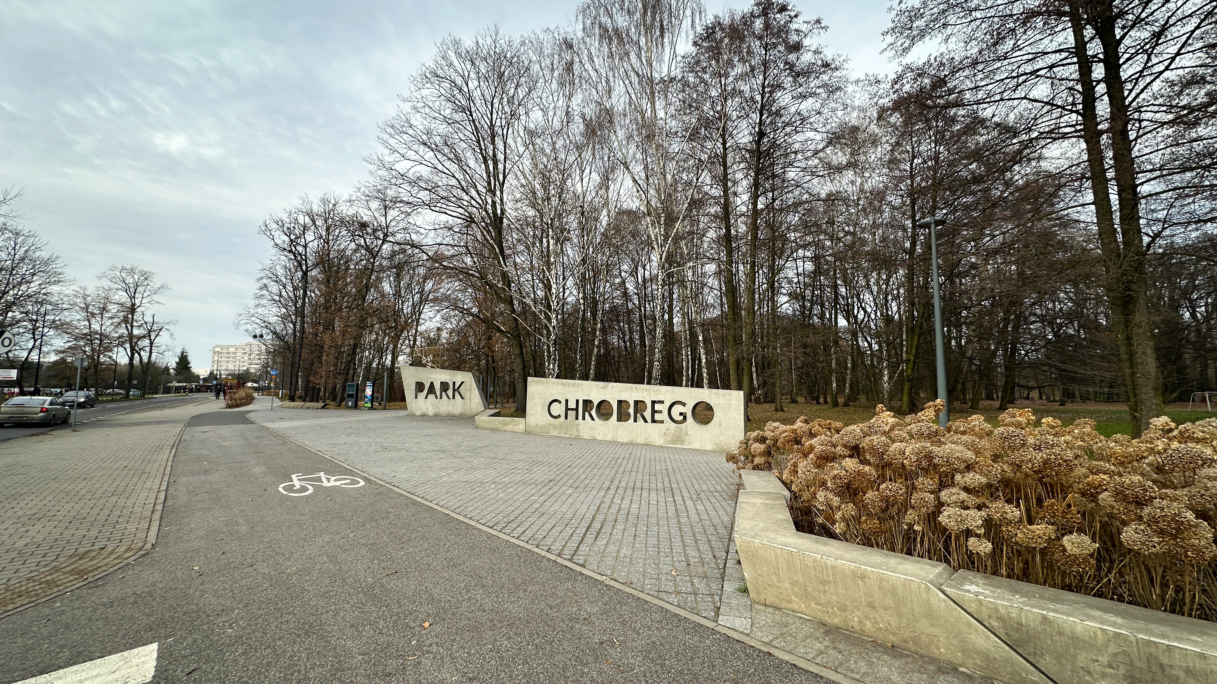 "Wejście do Parku Chrobrego oznaczone betonowymi konstrukcjami z dużym napisem 'PARK CHROBREGO'. W tle widoczne są drzewa bez liści, sugerujące porę jesienno-zimową. Po lewej stronie znajduje się ścieżka rowerowa, a po prawej – rabata z zaschniętymi rośli