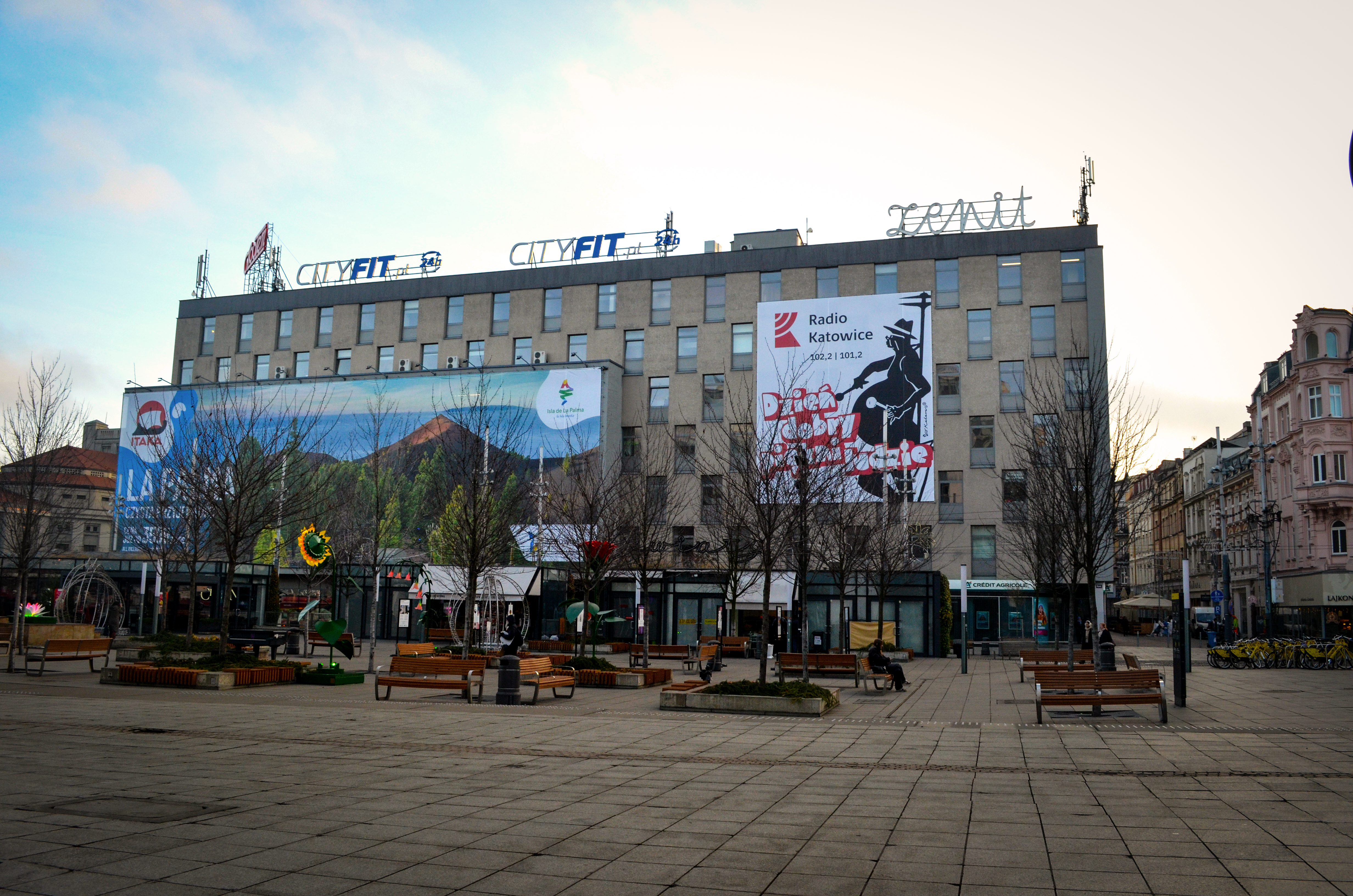 "Plac Kwiatowy w Katowicach z licznymi ławkami, drzewami bez liści i ozdobami kwiatowymi rozmieszczonymi na całej powierzchni placu. W tle znajduje się prostokątny budynek z reklamami i banerami, w tym z logo Radia Katowice oraz napisem 'Dzień Dobry w Rad